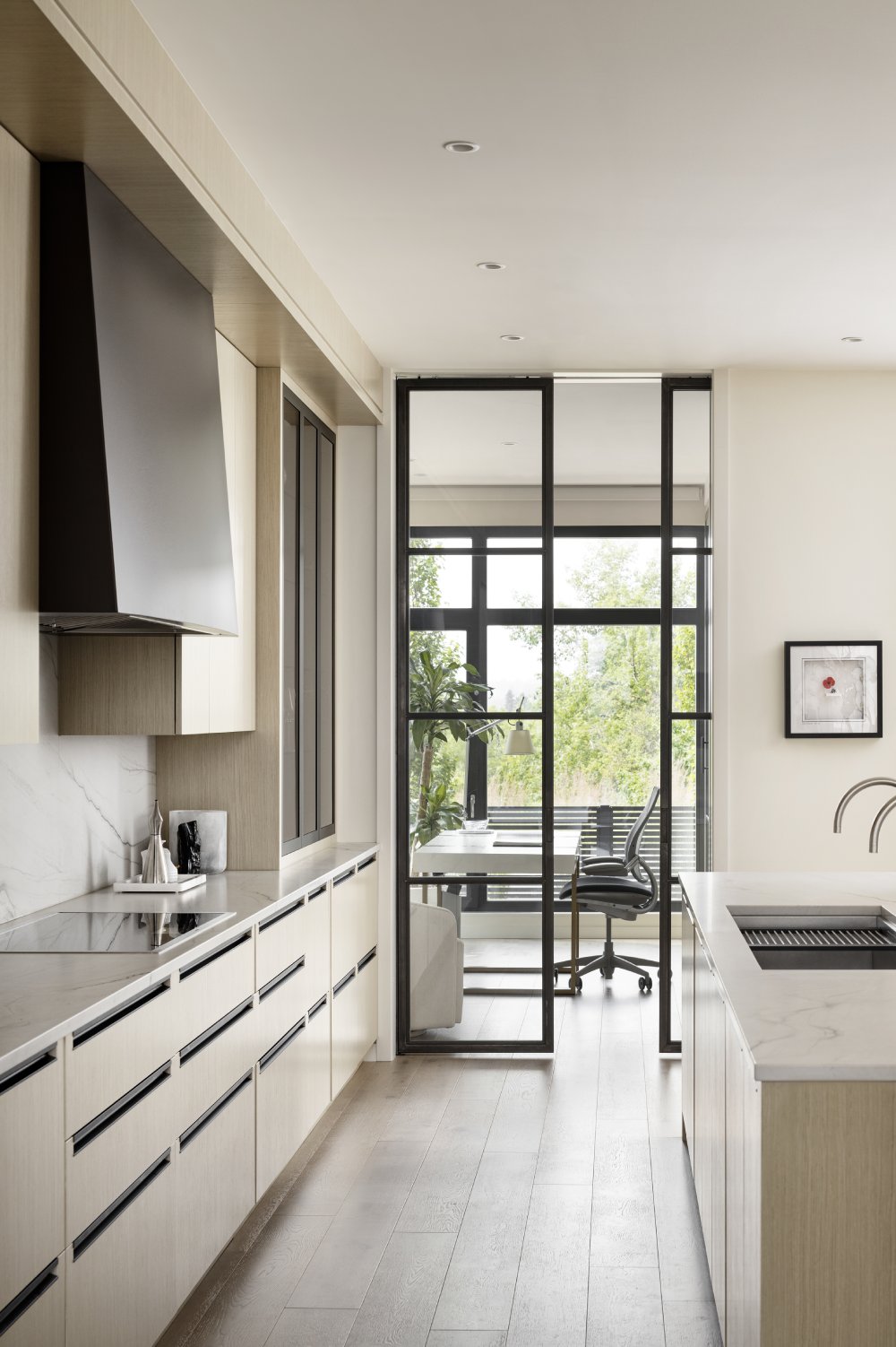 Looking through a beechwood cabinet kitchen at a home office with minimalist desk and roller chair in front of a window.