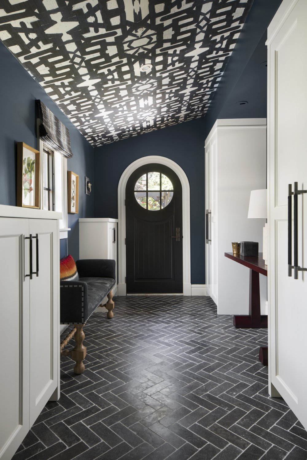 Hallway lined with white cabinetry looking at arched door with black herringbone tile floor and black and white patterned ceiling.
